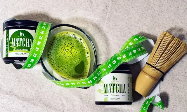 Zielony napój matcha w szklance, obok dwa słoiczki z napisem "MATCHA ORGANICZNA" i miarka krawiecka. Matcha co to? To zdrowy napój.