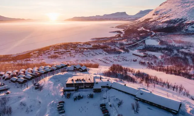 Vinterlandskap vid Kiruna med snötäckta fjäll, en frusen sjö och små stugor. Solen går ner och färgar himlen rosa.