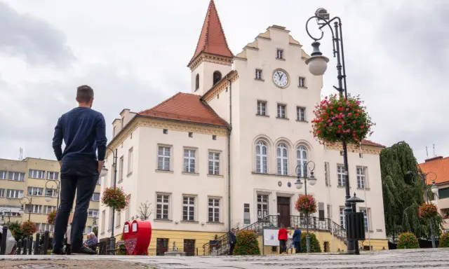 Mężczyzna podziwia ratusz w Trzebnicy, jedną z głównych atrakcji miasta. W tle czerwone serce i kwitnące kwiaty.