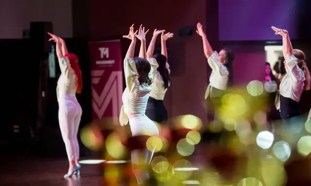 Tancerki w jasnych bluzkach i białych spodniach wykonują zmysłowy taniec. Ich dłonie unoszą się w powietrzu, tworząc dynamiczną choreografię. To piękny pokaz bachata solo.