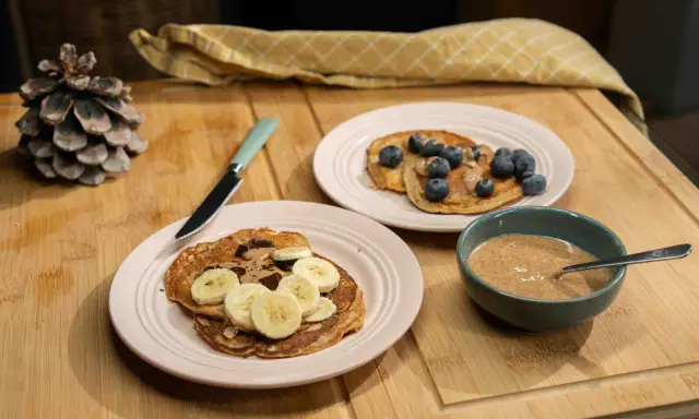 Pancakes ig bas farine complète, garnis de bananes, de chocolat et de myrtilles, avec un bol de beurre d'amande.