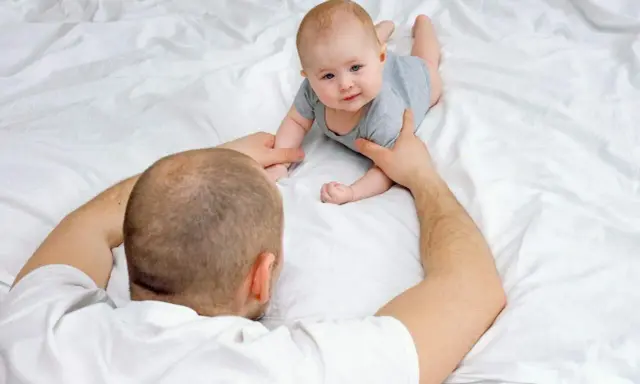 Tata pomaga dwumiesięcznemu dziecku obrócić się z brzucha na plecy. Maluch z zaciekawieniem patrzy na tatę.