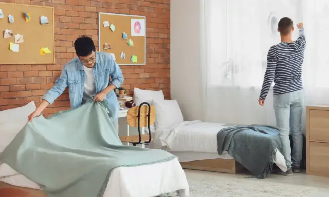 Dos jóvenes preparan su habitación de studea. Uno tiende una manta en la cama, el otro ajusta las cortinas.