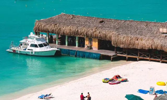 A boat docks at a thatched-roof beach bar on a sunny day. This scene embodies the welcoming spirit of Aruba, a destination known for being gay friendly.