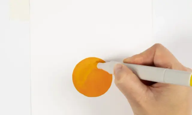 En hand håller en grå tuschpenna och målar en orange cirkel på vitt papper.