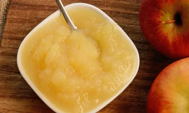 Frisches Apfelmus mit Schale, zubereitet im Thermomix, in einer weißen Schale auf Holzuntergrund. Daneben liegen zwei rote Äpfel.