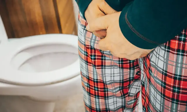 Osoba w piżamie z kratką przy toalecie, odczuwająca parcie na mocz przy cewniku.