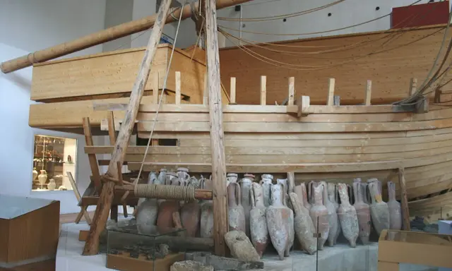 Amphores antiques alignées devant une reconstitution de navire en bois, rappelant le transport de vin par un ancien pinardier.
