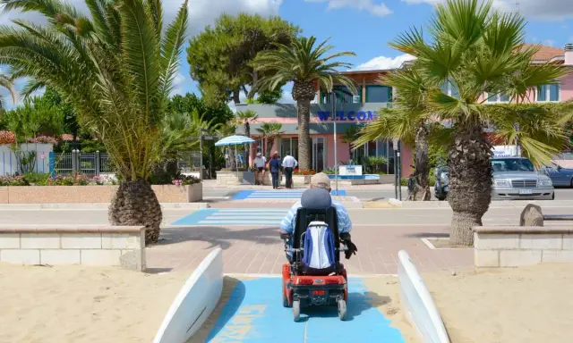 Accesso facilitato per tutti nei villaggi in Abruzzo sul mare. Un uomo in carrozzina elettrica percorre una passerella blu sulla sabbia, tra palme rigogliose e un edificio con la scritta "Welcome".