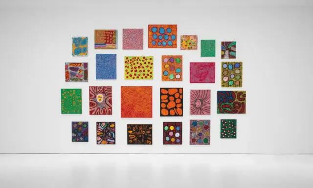 A vibrant collection of paintings by top visual artists, featuring bold colors and abstract patterns, adorns a white gallery wall.
