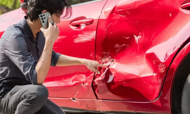 Hombre llama por teléfono señalando el grave golpe en la puerta de su coche rojo.