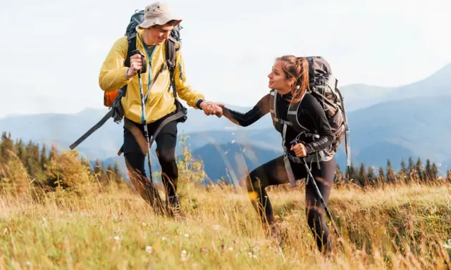 Coppia di escursionisti si aiutano a vicenda su un sentiero di montagna, con zaini e bastoncini da trekking.