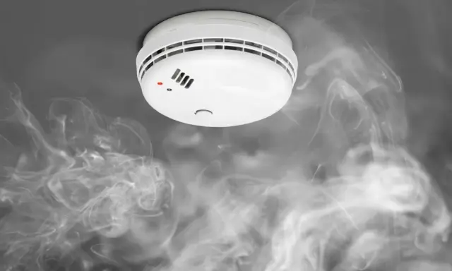 Biały czujnik dymu na suficie, otoczony kłębami dymu. Widać czerwoną diodę sygnalizującą alarm.