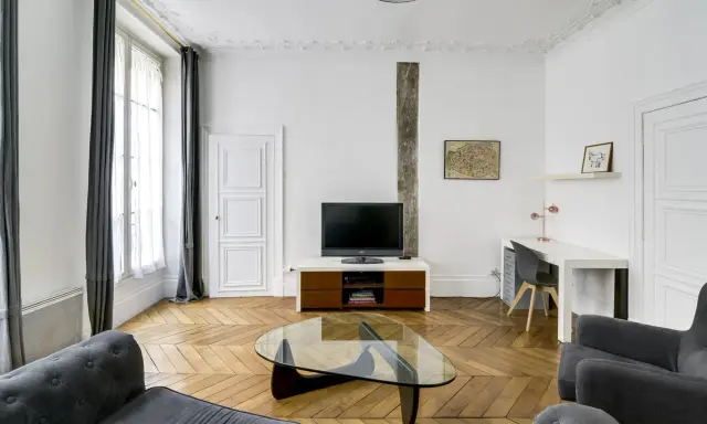 Salon moderne avec canapé gris, table basse en verre, bureau et mur blanc orné d'une carte de Paris.