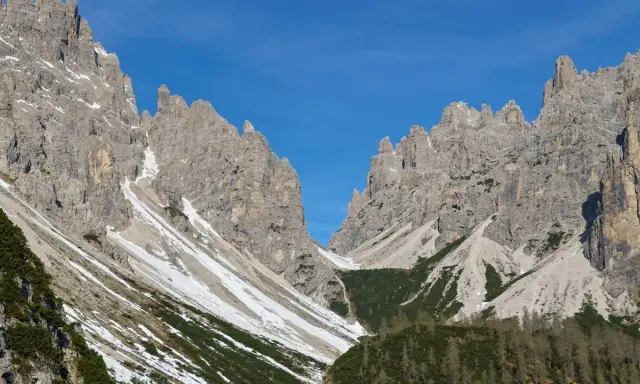 Vette rocciose imponenti sotto un cielo azzurro. Un sentiero scende verso il rifugio Giaf, tra pini e chiazze di neve.