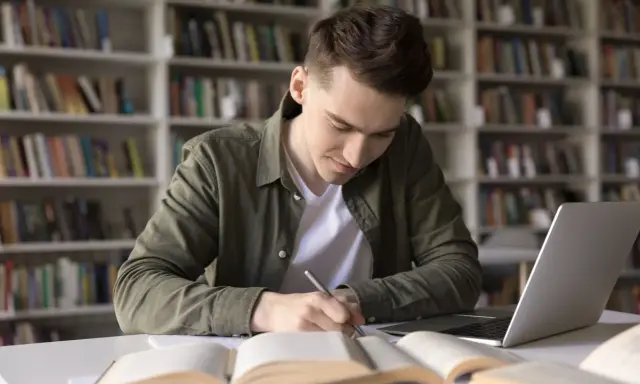 Młody mężczyzna w bibliotece pochyla się nad książkami i laptopem, pisząc notatki. Zastanawia się, czy technikum wlicza się do stażu pracy do emerytury.