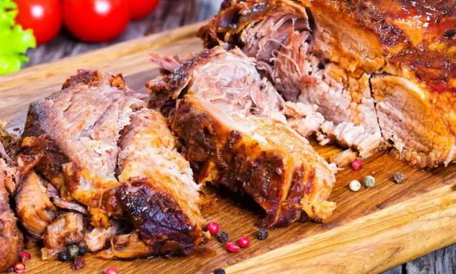 Une succulente épaule d'agneau rôtie, effilochée et servie sur une planche en bois, avec des tomates et de la laitue en arrière-plan.