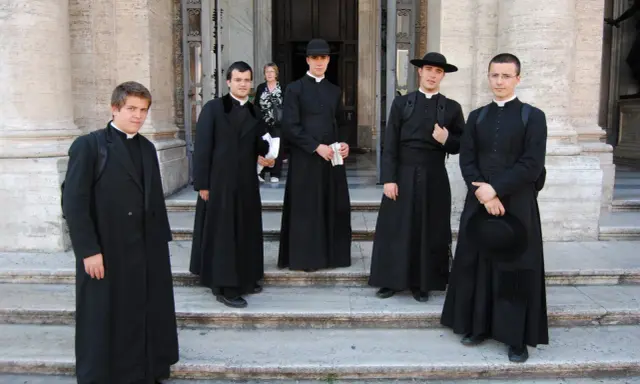 Cinque giovani seminaristi in abiti talari neri, alcuni con cappello, posano sulle scale di un edificio storico.