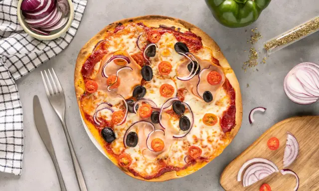 Gotowe ciasto na pizzę z szynką, oliwkami, pomidorkami i czerwoną cebulą. Obok sztućce i miseczka z cebulą.