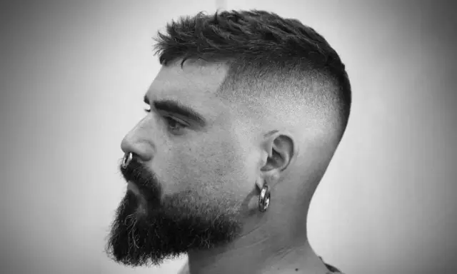 Mężczyzna z brodą i kolczykiem w nosie, z modnym, dłuższym buzz cut i cieniowanymi bokami.