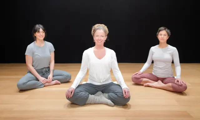Tre kvinnor sitter i lotusställning på ett trägolv. De deltar i en klass i traumaanpassad yoga.