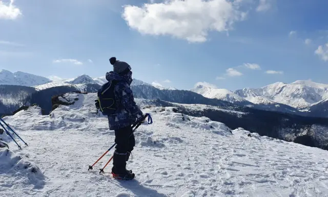 Chłopiec w zimowej kurtce i czapce z pomponem stoi na ośnieżonym szczycie, podziwiając wielki kopieniec gór. W rękach trzyma kijki trekkingowe.