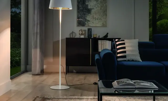 Biała, minimalistyczna lampa stojąca oświetla przytulny salon z granatową kanapą i szklanym stolikiem.