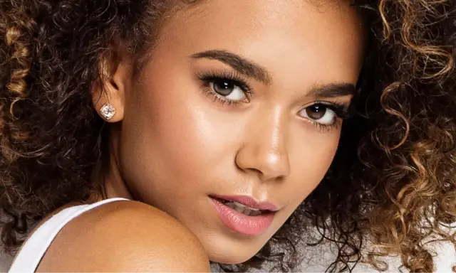 Un gros plan sur le visage d'une jeune femme aux cheveux bouclés, portant une boucle d'oreille étincelante. Ses **rehaucils avis** sont subtils, accentuant son regard.