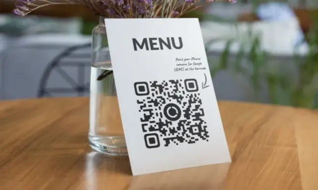 Un código QR de restaurante en una mesa de madera, junto a un jarrón con flores secas. Escanea para ver el menú.