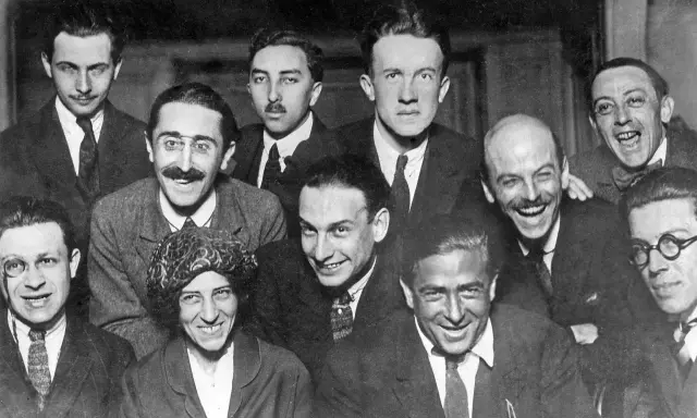 Un gruppo di uomini sorridenti, alcuni con baffi e occhiali, in una fotografia che evoca lo spirito irriverente del dadaismo.