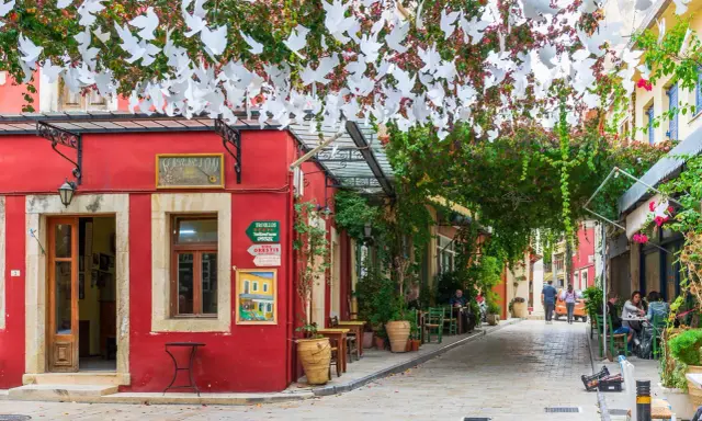 Ein rotes Gebäude mit weißen Tauben-Girlanden über einer Gasse in Chania, Kreta. Tische und Stühle laden zum Verweilen ein.