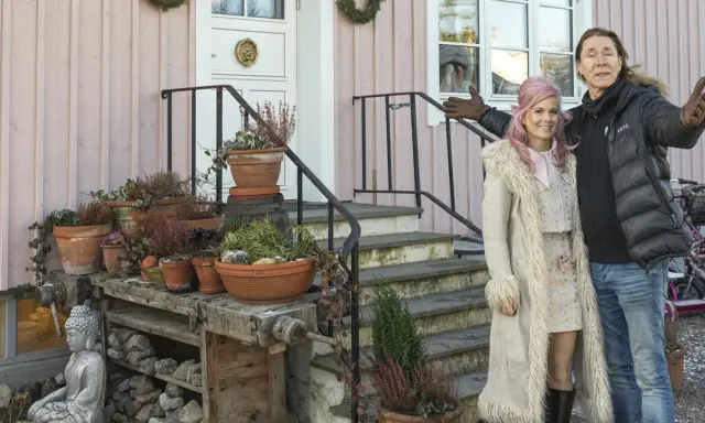 Melinda Jacobs och E-Type poserar glatt framför ett rosa hus med många krukor och en Buddha-staty.