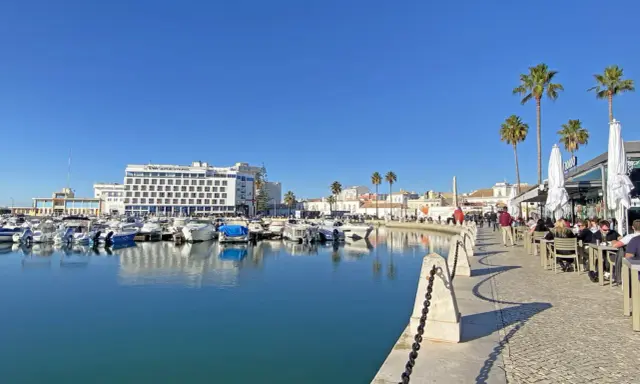 Boote im Hafen von Faro, Portugal, spiegeln sich im ruhigen Wasser. Palmen säumen die Promenade, wo Menschen in Cafés sitzen.