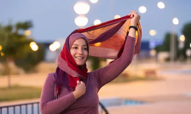 Lächelnde muslimische Frau mit buntem Hijab und Schal, der im Wind weht. Ein Bild, das die Vielfalt und Schönheit islamischer Kultur zeigt.