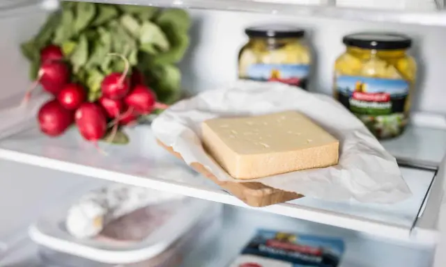 Ein Stück Käse liegt auf einem Holzbrett, daneben Radieschen und Gläser mit eingelegtem Gemüse. Verschiedene Käse sorten im Kühlschrank.