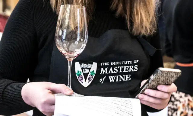 Eine Person mit einer Schürze des "Institute of Masters of Wine" hält ein Weinglas und ein Smartphone.