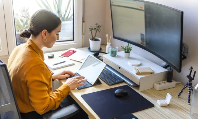 Mujer trabajando en un escritorio ordenado, con portátil, monitor curvo y plantas. Un ejemplo de como organizar tu escritorio para ser productivo.