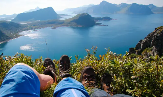 Fötter i vandringskängor vilar på en gräsbevuxen bergssluttning med en fantastisk utsikt över Lofoten. En perfekt dag för lofoten vandring.