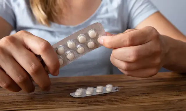 Dłonie trzymają opakowanie z tabletkami, które mogą być lekami z serotoniną. Na drewnianym blacie leży drugie, częściowo zużyte opakowanie.