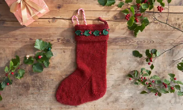 Röd stickad julstrumpa med grön misteldekoration och polkagrisar. En present och julgranspynt ligger på träbordet.