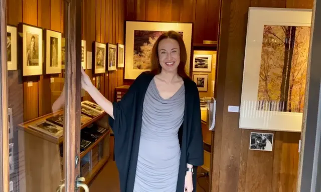 A woman smiles, opening the door to a photography west gallery filled with framed prints and display cases of artwork.
