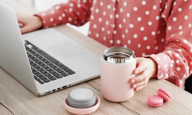 Kobieta przy laptopie trzyma kubek termiczny, obok leży nakrętka. Dowiedz się, jak wyczyścić nakrętkę od kubka termicznego, by napoje dłużej smakowały świeżo.