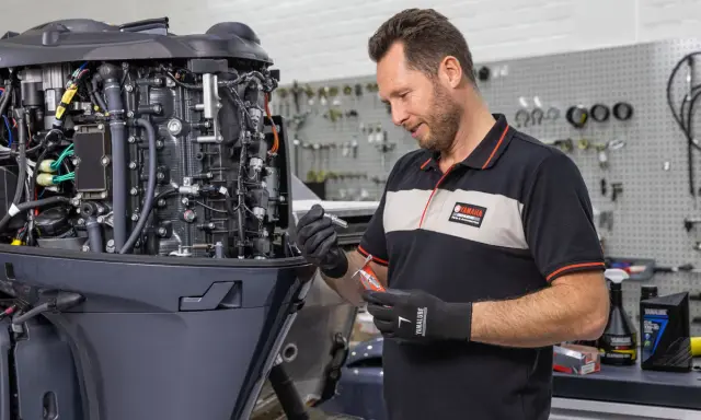 Un mécanicien effectue l'entretien d'un moteur hors-bord 4 temps Yamaha, tenant une bougie et un outil.