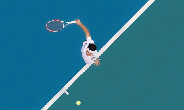 A tennis player in mid-serve, racket raised, on a blue and green court. The white lines of the court are like brad walls, guiding the game.