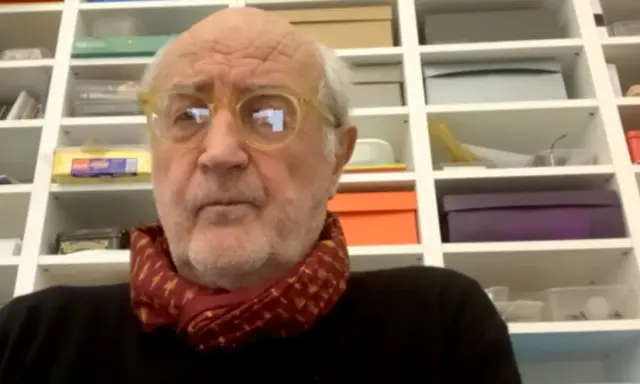 Ritratto di Francesco Pecoraro con occhiali gialli e sciarpa rossa, davanti a una libreria piena di scatole.