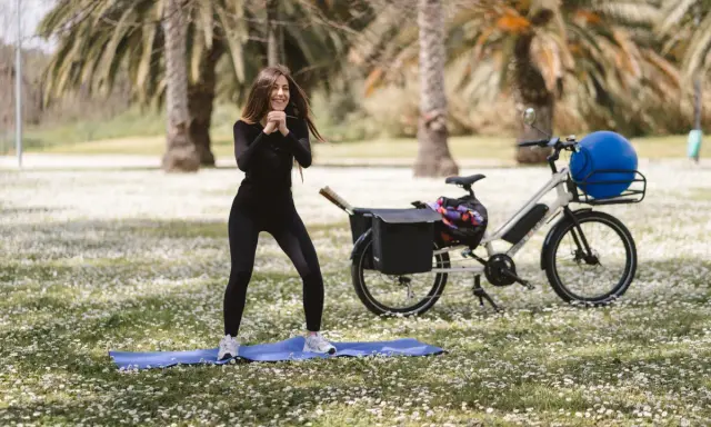 Donna sorridente fa esercizi all'aperto, accanto a una bicicletta per dimagrire con un grande pallone blu nel cestino.