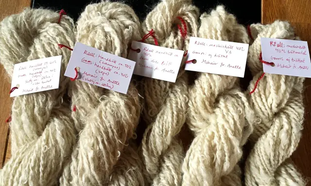 Buntar av ljusbeige garn, troligen mohair ull, med handskrivna etiketter som beskriver tvättmetoder och ursprung.