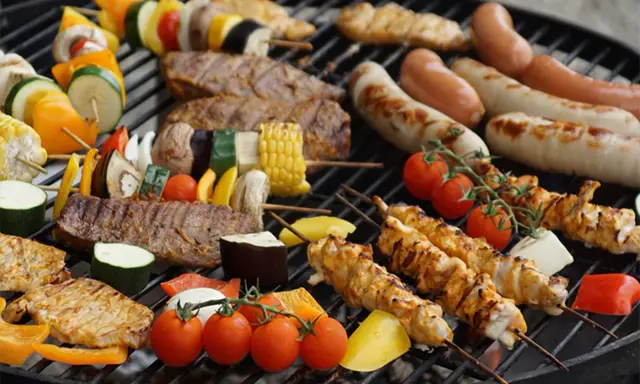 Una parrillada de que es bbq: brochetas de pollo y verduras, salchichas, bistecs y mazorcas de maíz asándose en una parrilla.
