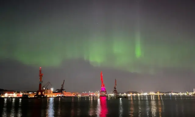 Norrsken över Göteborgs hamn. Kranar lyser upp natten medan himlen dansar i grönt.