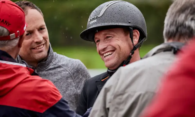 Michael Jung lacht in die Kamera, während er mit anderen Personen spricht. Er trägt einen schwarzen Reithelm mit Regentropfen.